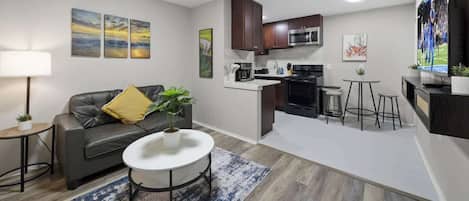 A modern living room with a grey sofa, a round coffee table, and a kitchen area with dark cabinets and a microwave.