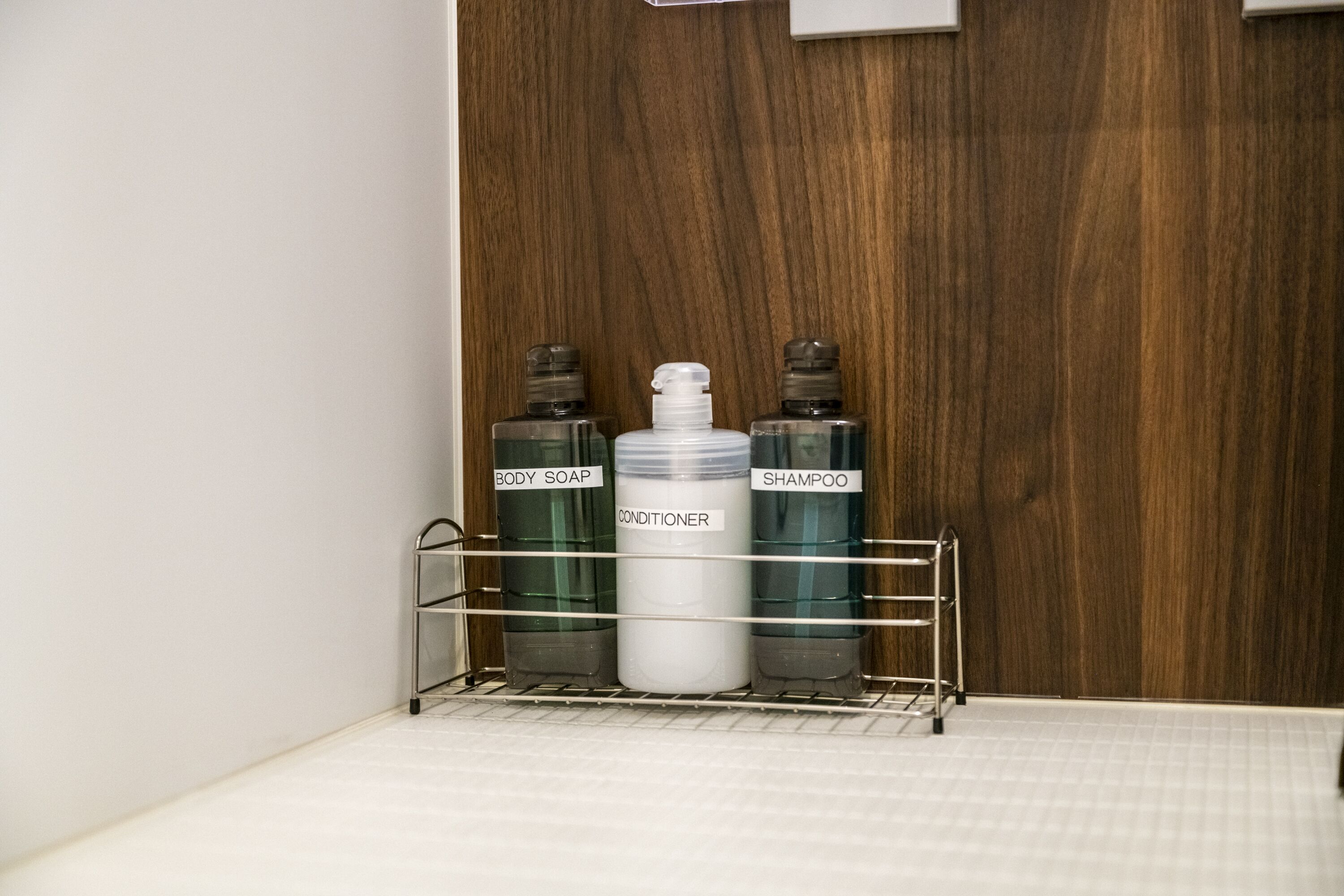 A bathroom with a wooden wall, a metal shelf holding bottles labeled "BODY SOAP," "CONDITIONER," and "SHAMPOO."