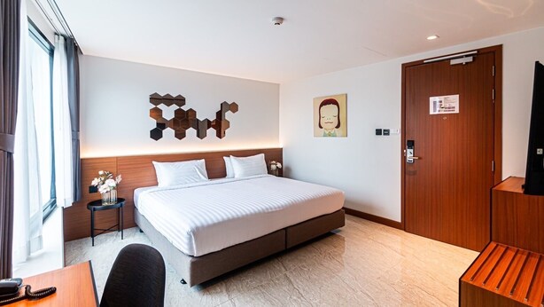 A modern hotel room with a large bed, a desk with a telephone, a small side table with a vase, a wooden wardrobe, and a wall-mounted artwork.