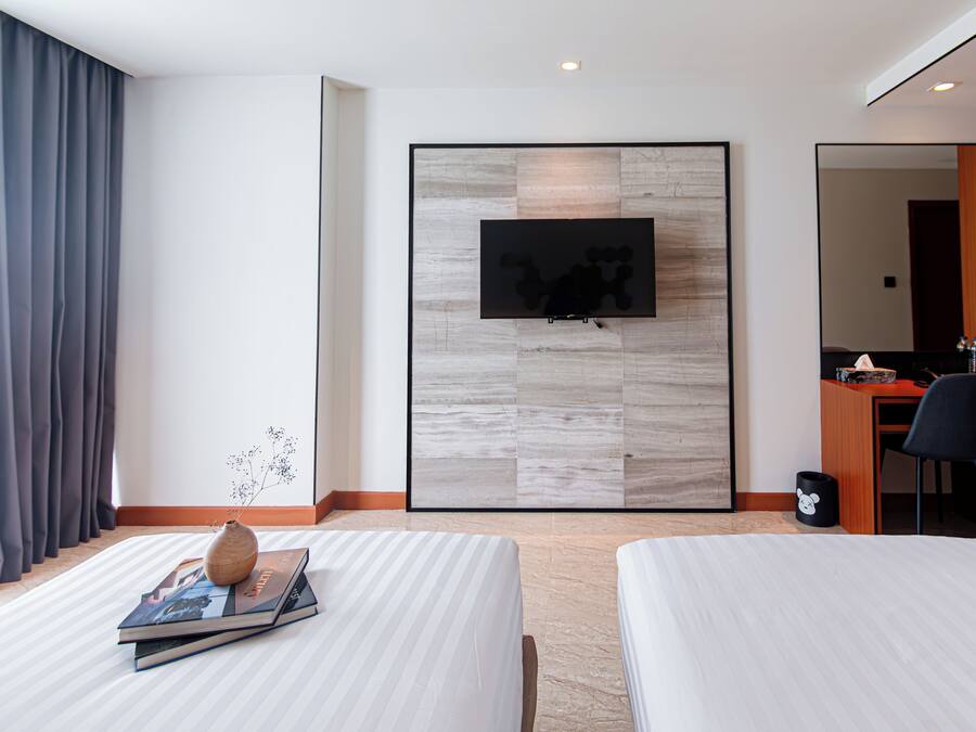 A hotel room with two beds, a TV mounted on a wooden panel wall, a desk with a chair, and a small trash bin.