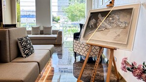 A living room with a sofa, an easel with a painting, and a view of the outdoors through large windows. - Frame Hotel Bangkok (Bangkok)