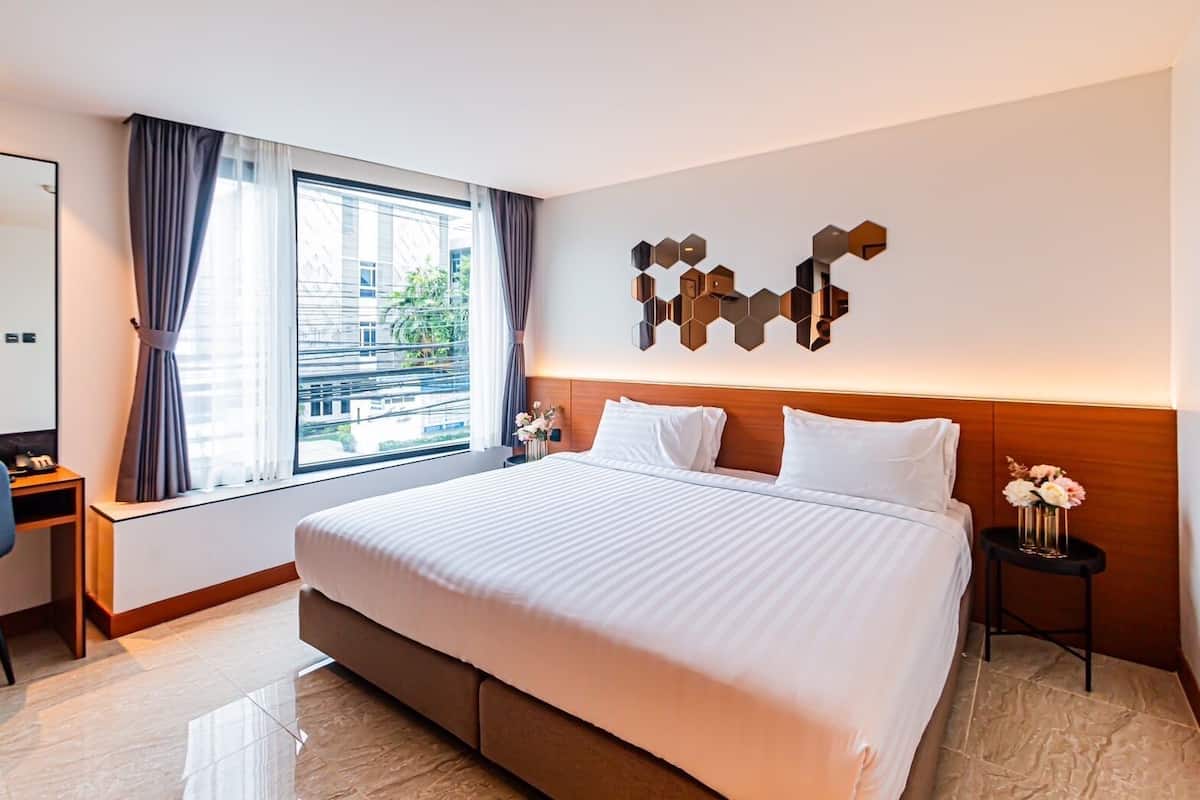 A hotel room with a large bed, a desk, a chair, a window with curtains, and a wall-mounted mirror.