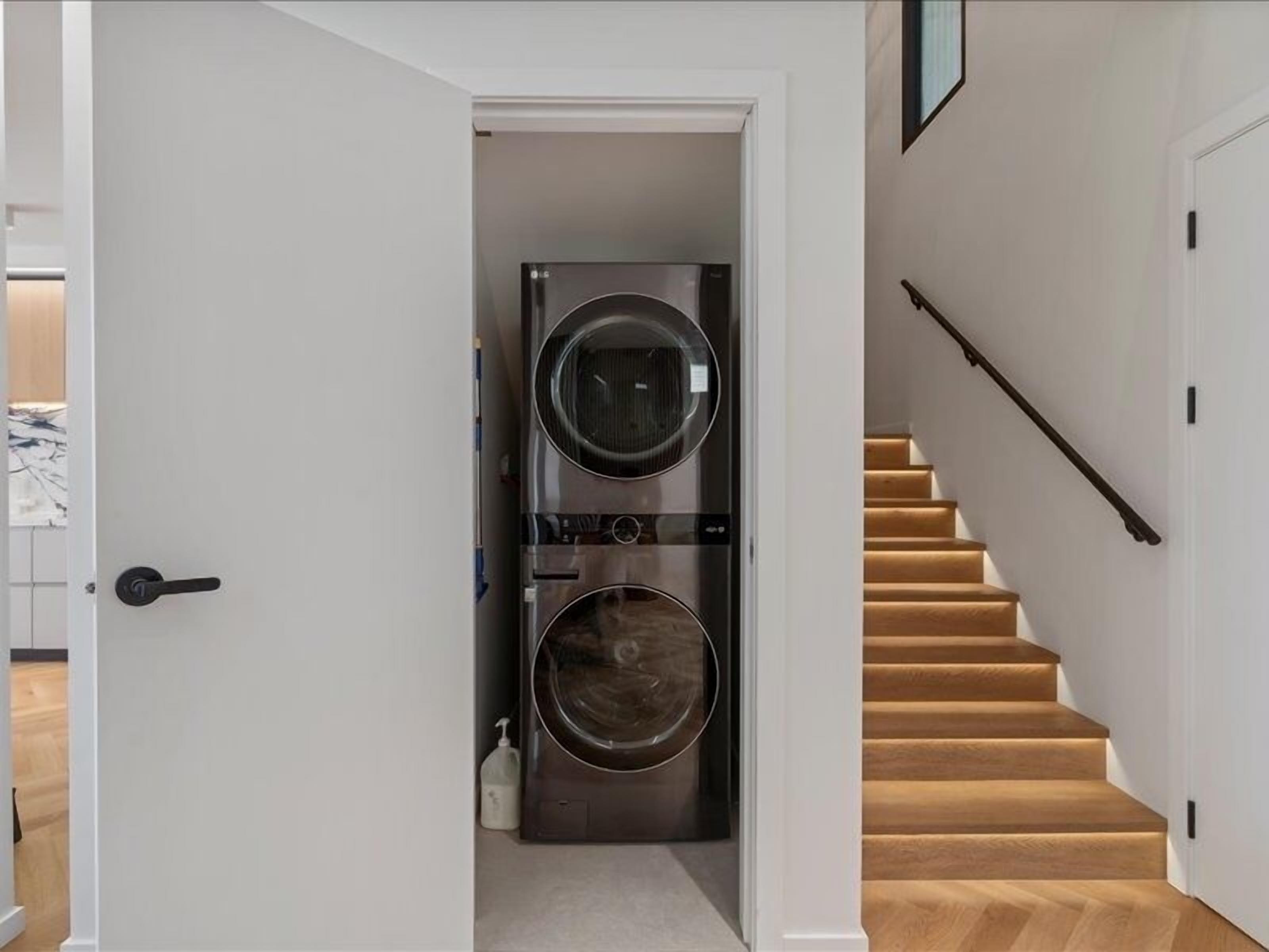 A laundry room with two stacked front-loading washing machines, a white door, and a wooden staircase with a handrail.