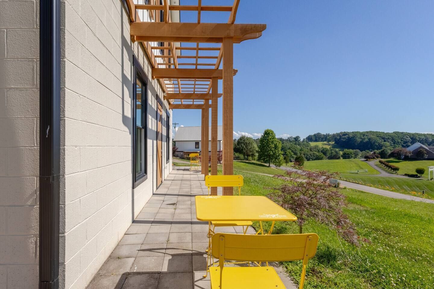 A building with a covered outdoor area, yellow tables and chairs, and a view of a green landscape.