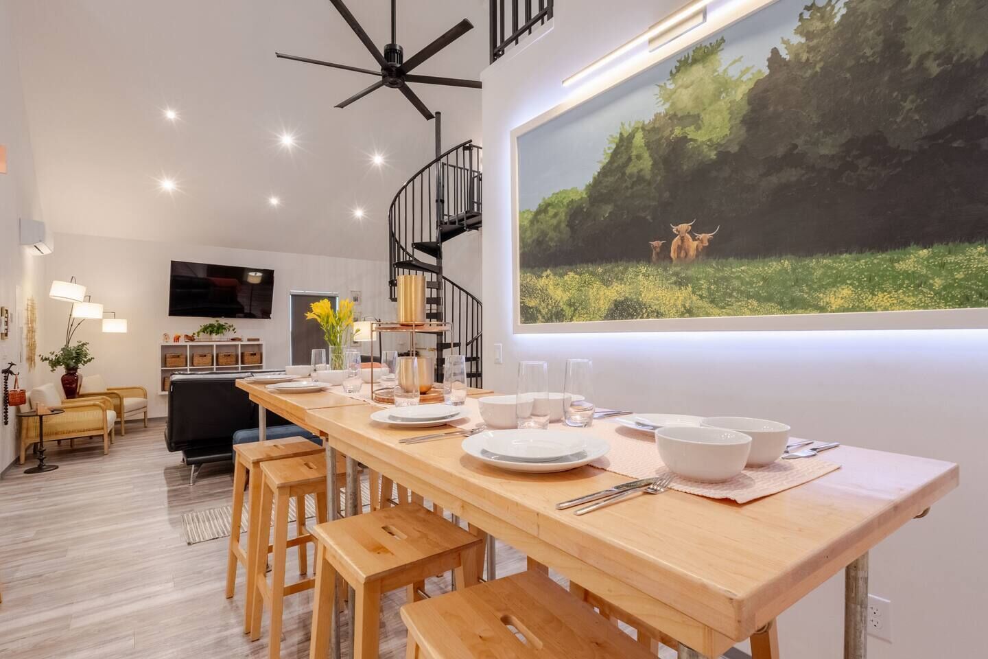 A modern dining area with a wooden table set for four, a black spiral staircase, and a large window with a nature scene.