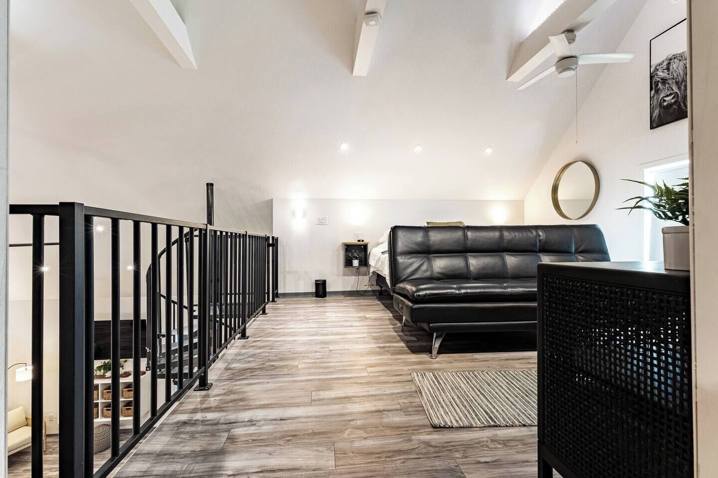 A modern interior with a black leather sofa, a wooden floor, a black railing, and a ceiling light fixture.