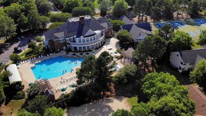 An aerial view of a property with a large swimming pool surrounded by lounge chairs and umbrellas, a tennis court, and a large house with a curved roof.