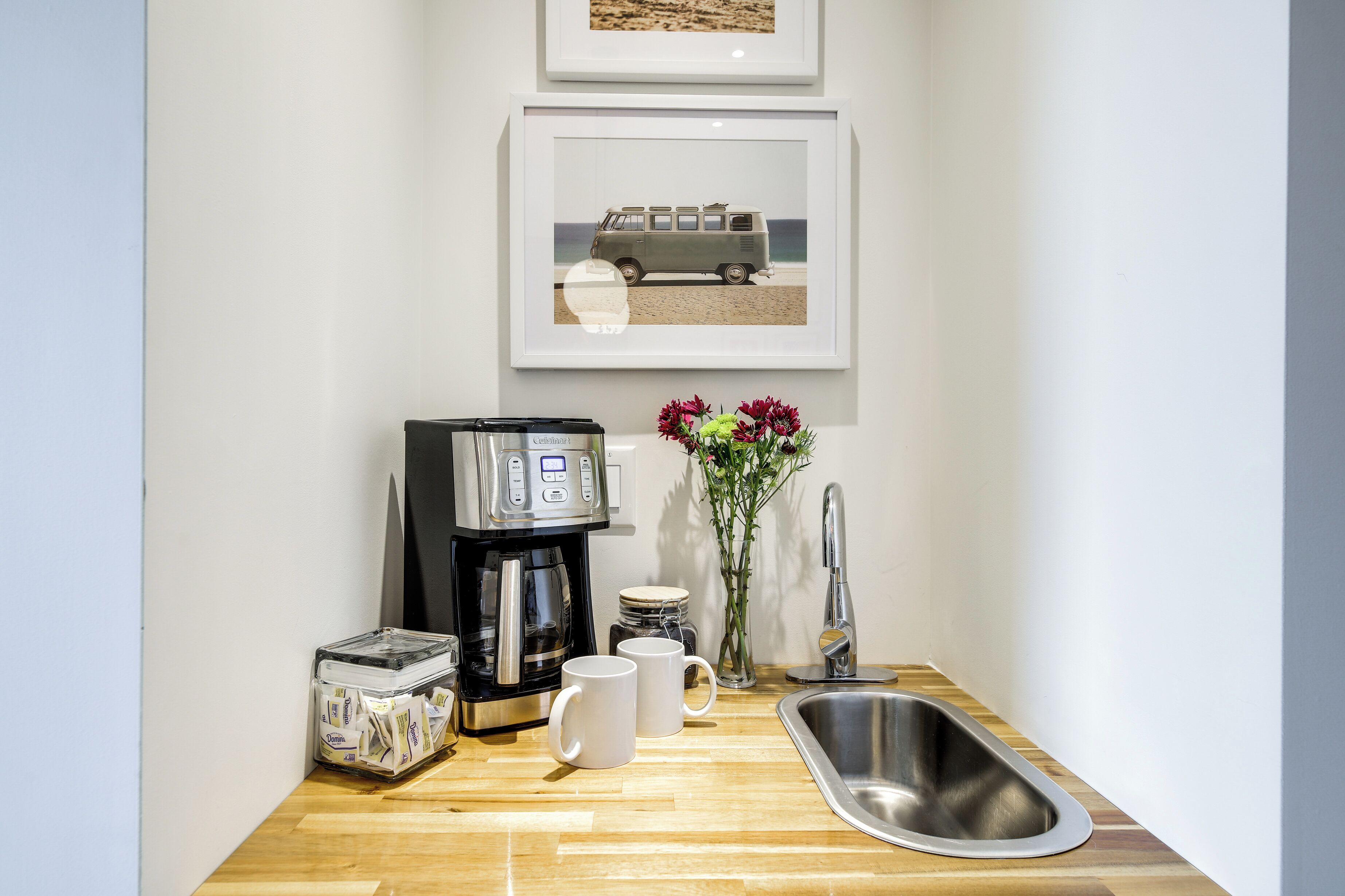 A kitchenette with a coffee maker, two mugs, a sink, and a vase with flowers.