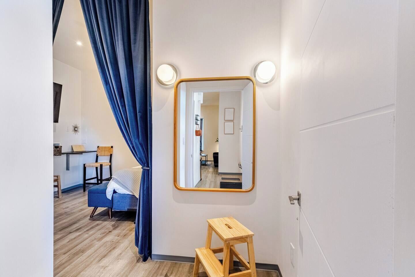 Una habitación de hotel con una cama, un taburete de madera, un espejo y una cortina.