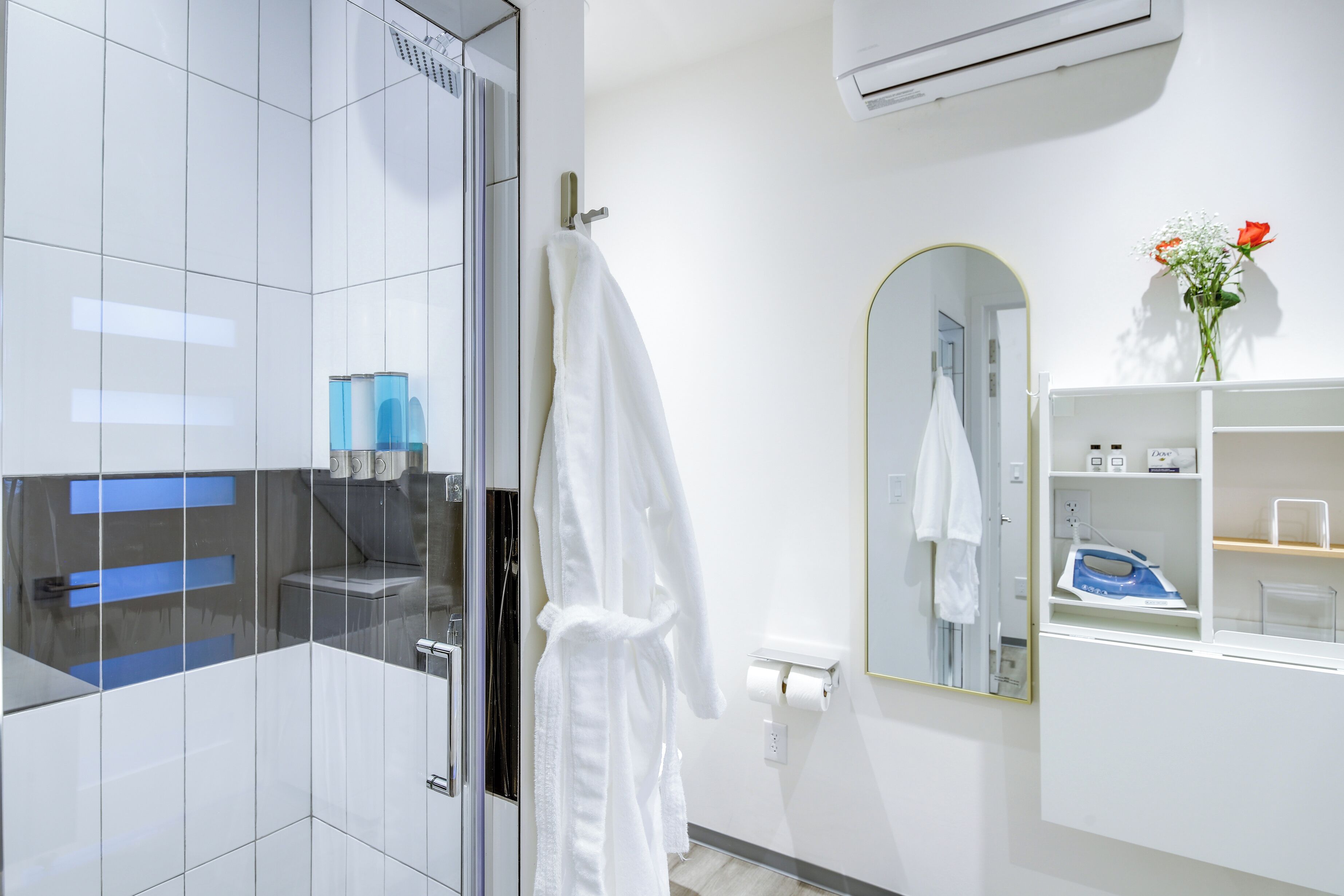 A modern bathroom with a glass shower enclosure, a white robe hanging on a rack, a mirror with a gold frame, and a shelf with toiletries.