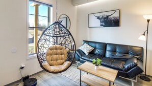 A modern living room with a hanging chair, a black leather sofa, a wooden coffee table, and a framed artwork on the wall.