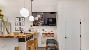 A modern kitchen with a wooden island, pendant lights, a dining area with wooden chairs, and a TV mounted on the wall.