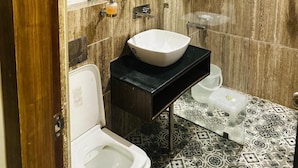 A bathroom with a marble wall, a white sink, a toilet, and a shower area. - Hotel Fortune Ganga (Haridwar)