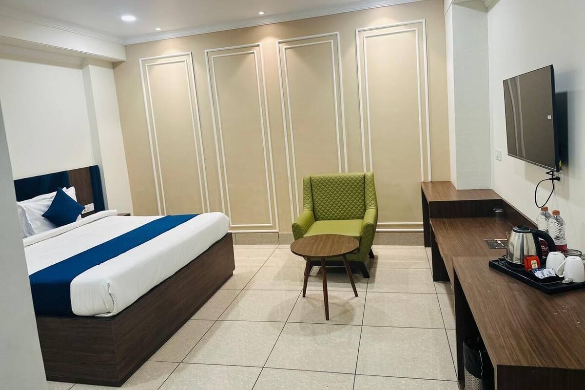 A hotel room with a bed, a green armchair, a wooden desk, a television, and a coffee station.