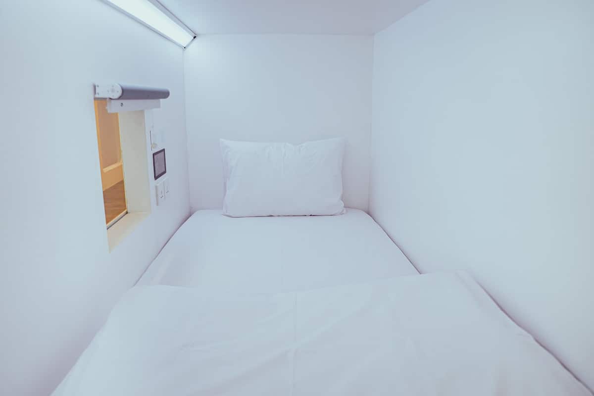 A small, white, single-bed room with a simple layout, a small window, and a power outlet on the wall.
