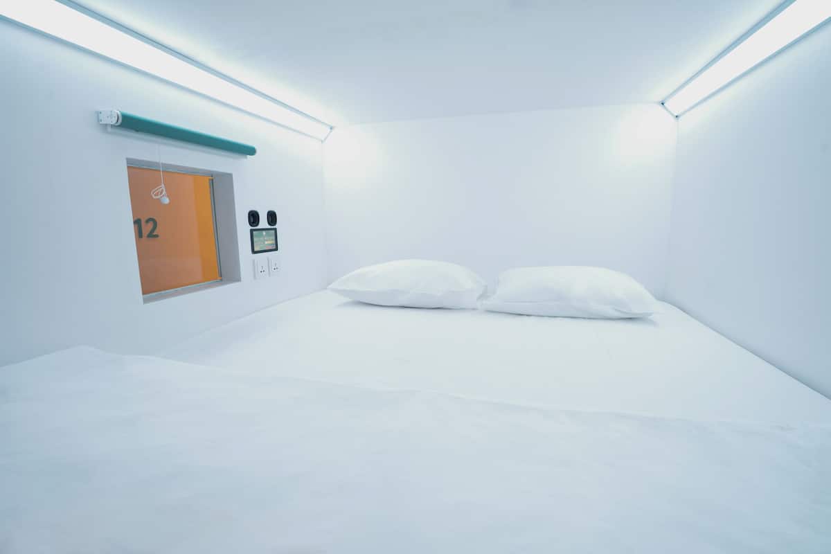A minimalist hotel room with a bed, a control panel, and a door marked with the number 12.