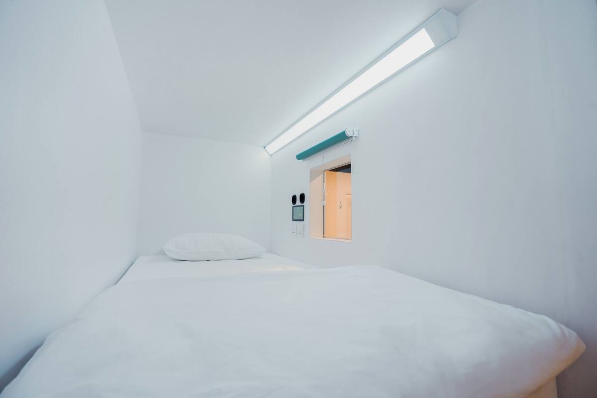 A minimalist room with a bed, a small window, and a wall-mounted control panel.