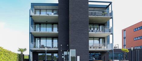 A modern multi-story building with balconies, a central entrance, and a paved ground area.