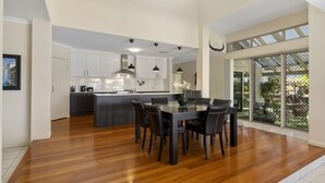 A modern kitchen with a central island, a dining area with a glass table, and a large sliding glass door leading to a balcony. - Bribie Bliss: Luxe Canal Living (BANKSIA BEACH)