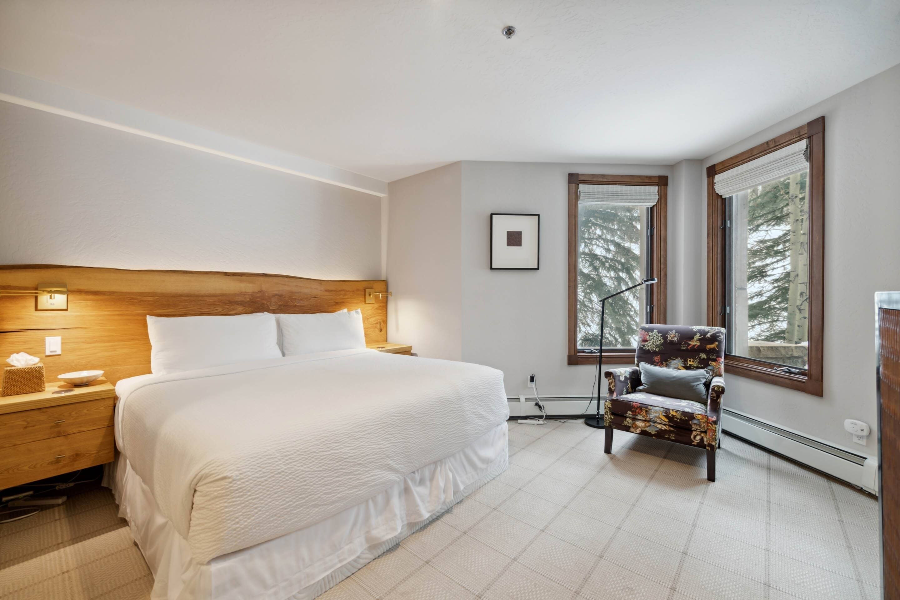 Un dormitorio con una cama grande, cabecera de madera, mesitas de noche, una silla y ventanas amplias con persianas.