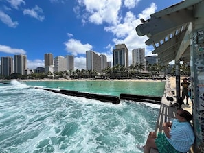 A beach with waves crashing onto the shore, a city skyline in the background, and a pier with people on it.