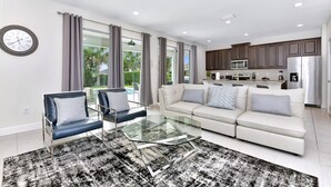 A modern living room with a glass coffee table, two armchairs, a sofa, and a kitchen in the background. - Cheerful Home near Disney with Private Pool, Themed Rooms and Waterpark &. (Kissimmee)