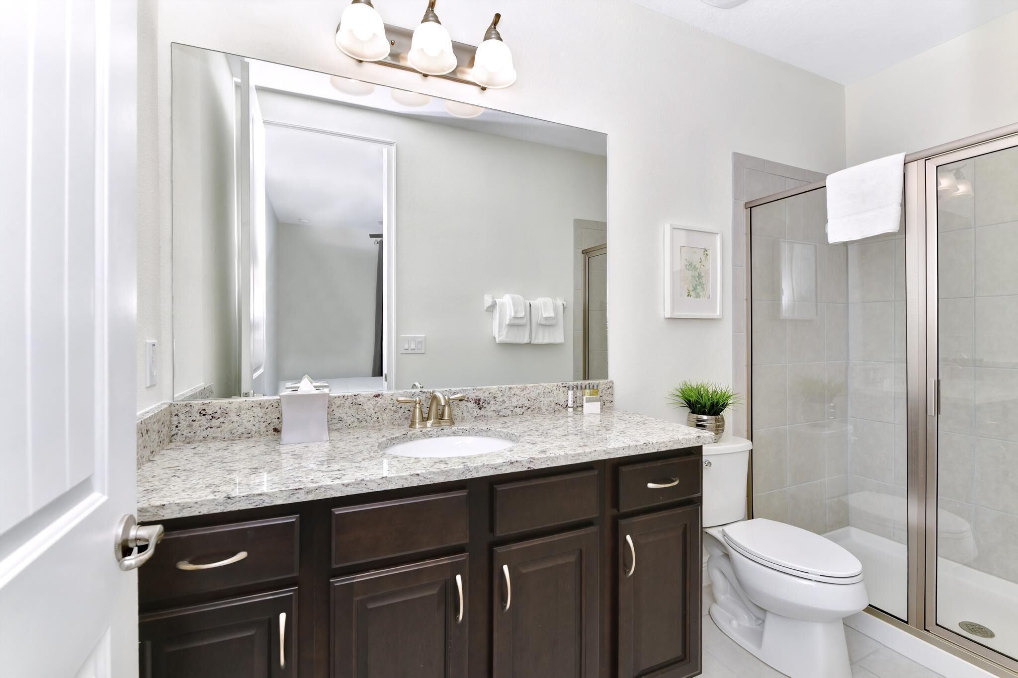 A modern bathroom with a granite countertop, a large mirror, a white sink, dark wooden cabinets, a white toilet, and a glass shower enclosure.
