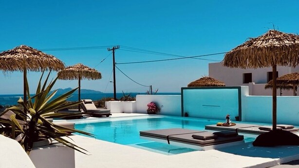 A pool area with lounge chairs and umbrellas, a clear blue sky, and a view of the sea. - The Vine Suites (Santorini)