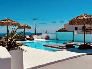 Zona de piscina con sillones de descanso y sombrillas, cielo azul despejado y vista al mar.
