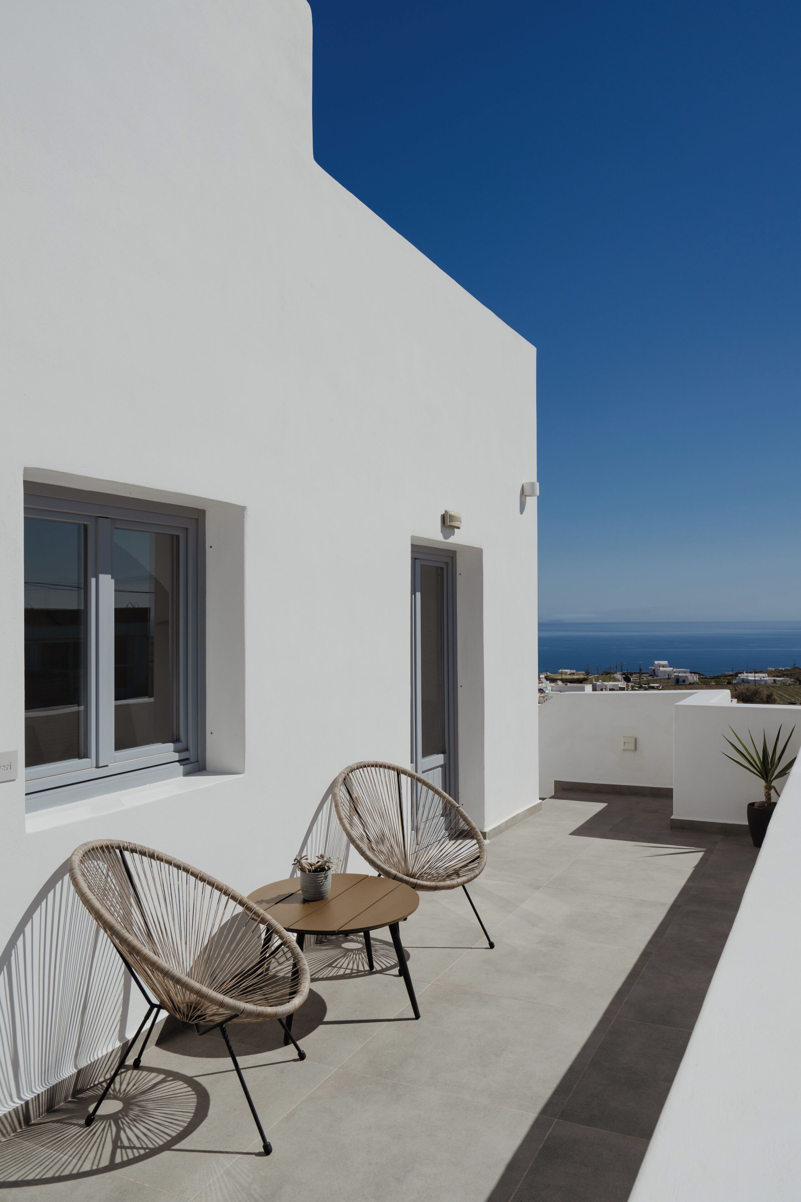 A modern outdoor balcony with two wicker chairs and a small table, overlooking a coastal view.