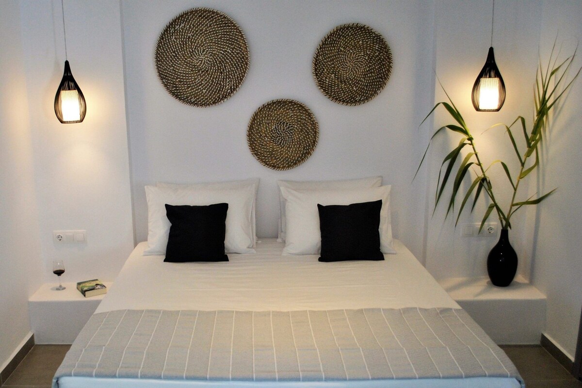 Un dormitorio ordenado con una cama, dos mesitas de noche, una lámpara de noche, un adorno en la pared y una planta en maceta.