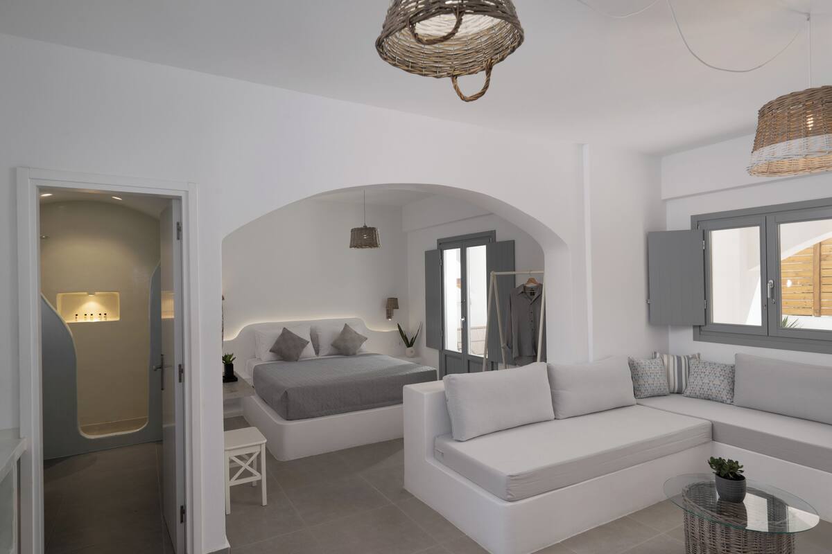 Un dormitorio moderno y minimalista con una cama, un sofá, una mesita y una silla de mimbre.