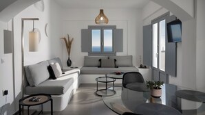 A modern living room with a sofa, a coffee table, a TV, and a window with a view. - The Vine Suites (Santorini)