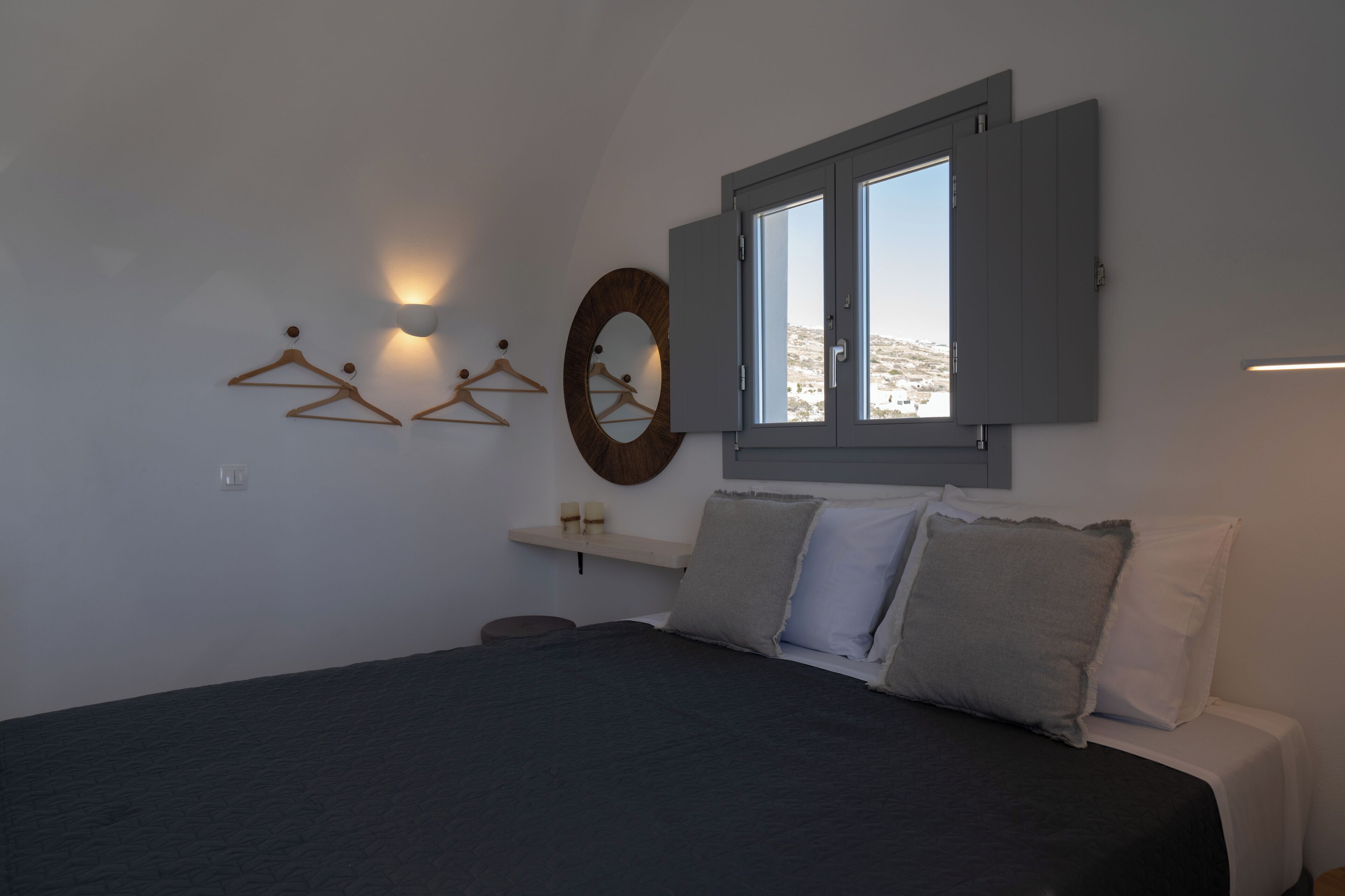Un dormitorio con una cama, una ventana con contraventanas abiertas, lámparas fijadas a la pared y un espejo circular.