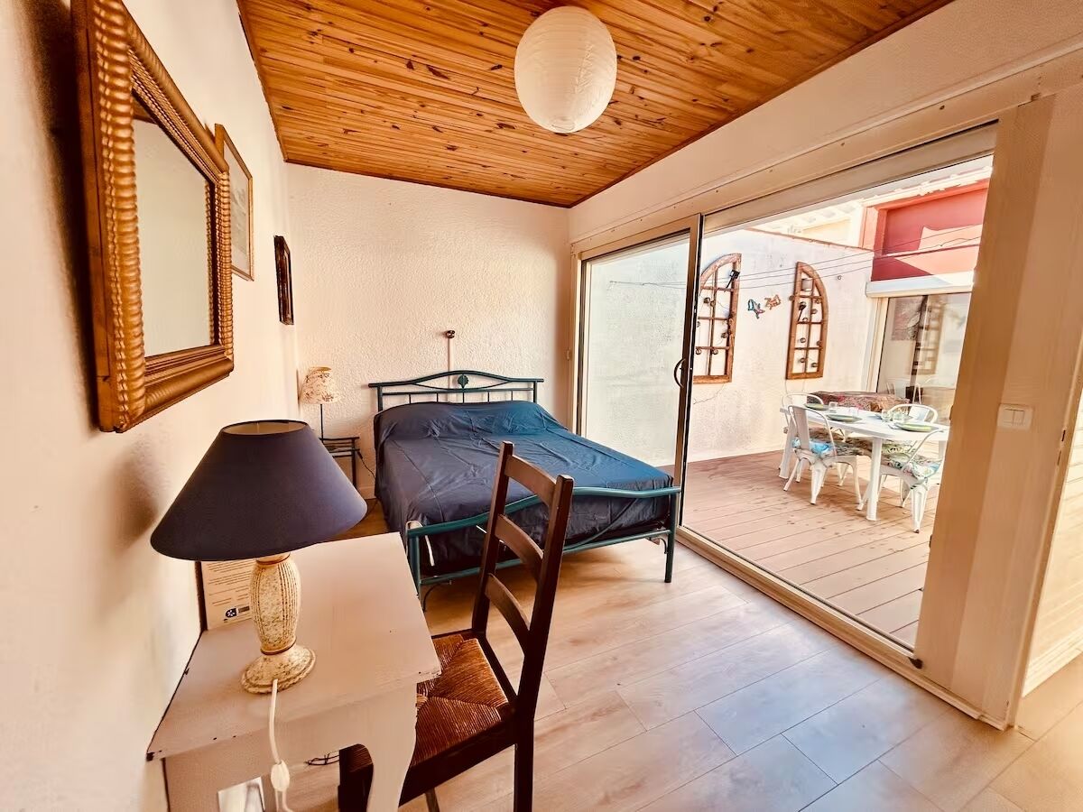 A bedroom with a bed, a chair, a desk with a lamp, and a sliding glass door leading to an outdoor area with a table and chairs.
