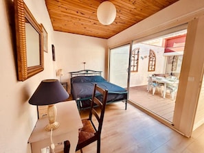 Un dormitorio con una cama, una silla, un escritorio con lámpara y un ventanal corredero que da a un espacio exterior con mesa y sillas.