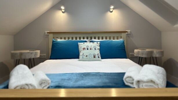 Ein Schlafzimmer mit geneigter Decke, einem Bett mit blauer Bettwäsche, einem Zierkissen, zwei Nachttischen und gerollten Handtüchern auf dem Bett.