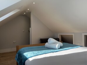 A bedroom with a sloped ceiling, a bed with a blue bedspread, and two rolled towels on the bed.