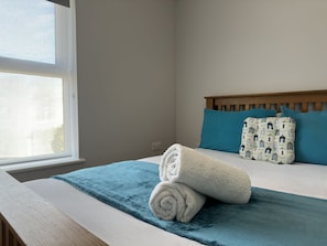 Cama bem arrumada com manta azul e toalhas brancas dobradas, cabeceira de madeira e dois travesseiros estampados.