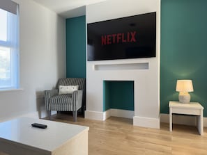 A modern living room with a flat-screen TV mounted above a fireplace, a striped armchair, a small side table with a lamp, and a remote control on a coffee table.