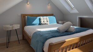 Un dormitorio con techo inclinado, tragaluz, cama con manta azul y mesita de noche con lámpara.