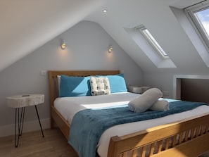 A bedroom with a sloped ceiling, a skylight, a bed with a blue blanket, and a bedside table with a lamp.