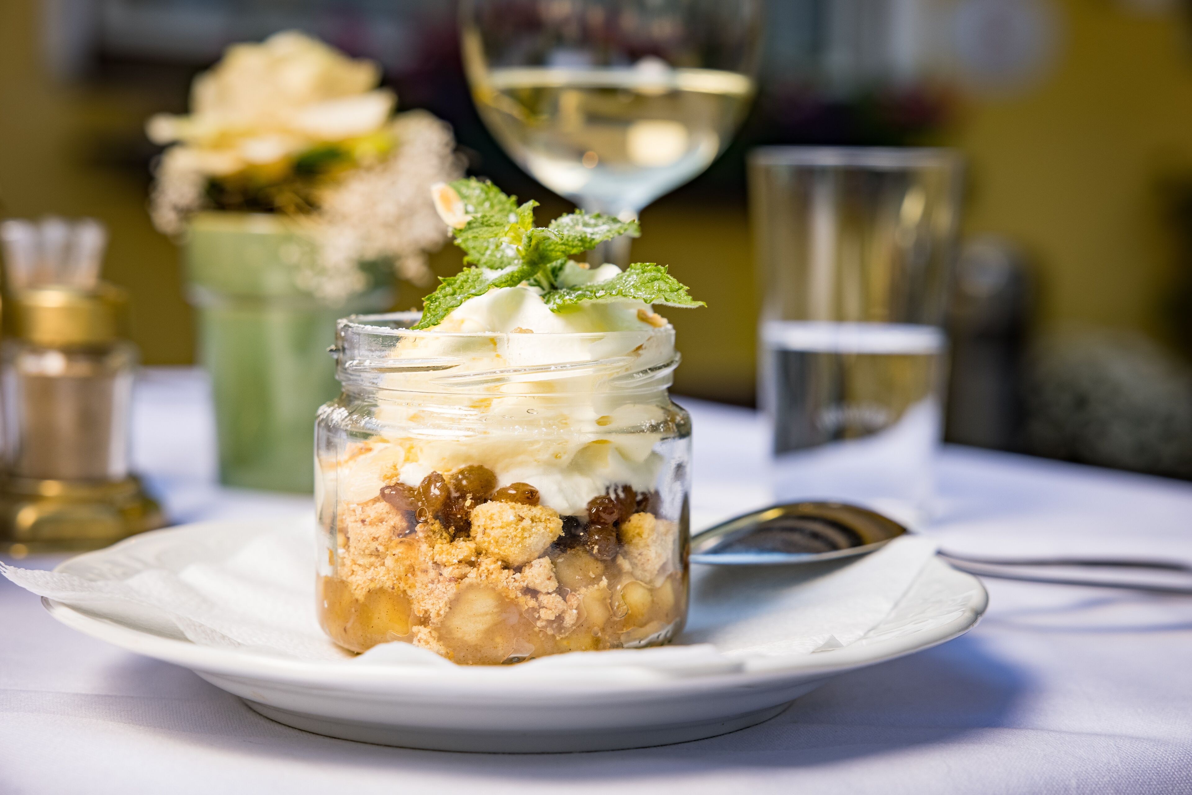 Un postre en un tarro sobre un plato blanco, con una copa de vino blanco y un vaso de agua al fondo.