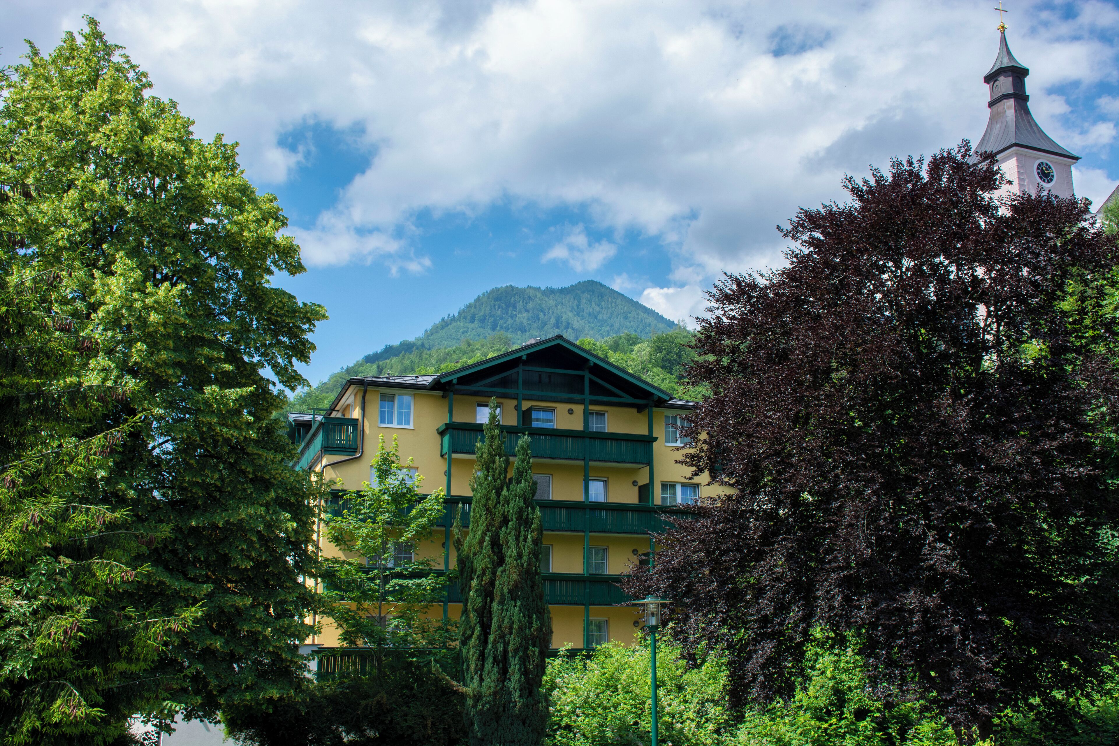 Un edificio de varios pisos con balcones, rodeado de exuberante vegetación y una montaña al fondo.