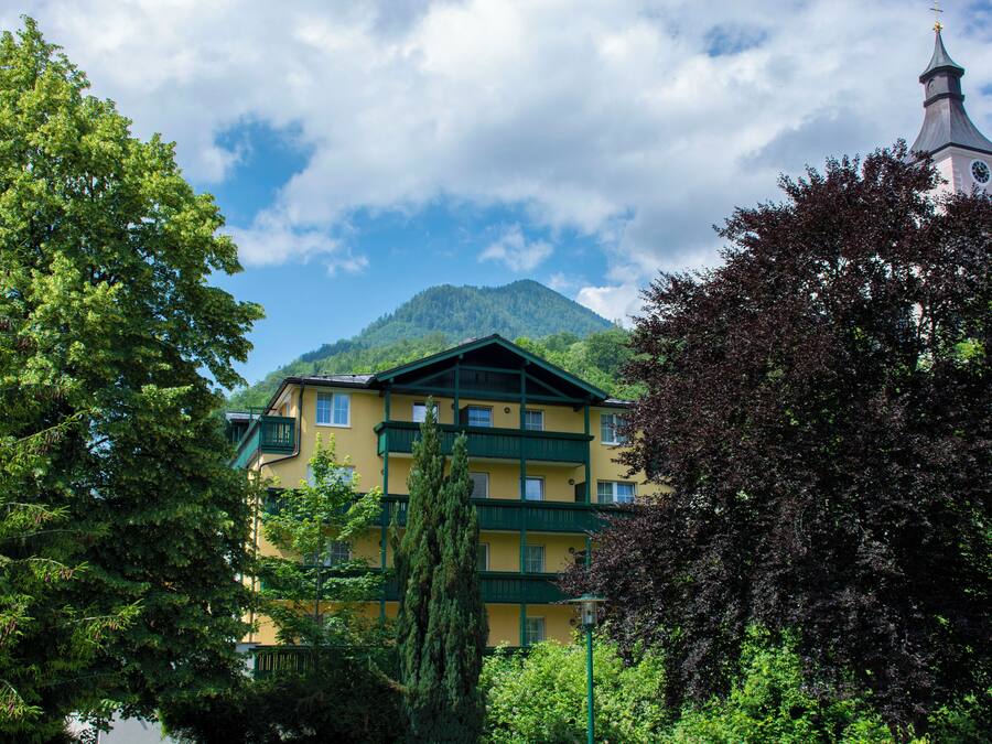 Un edificio de varios pisos con balcones, rodeado de exuberante vegetación y una montaña al fondo.