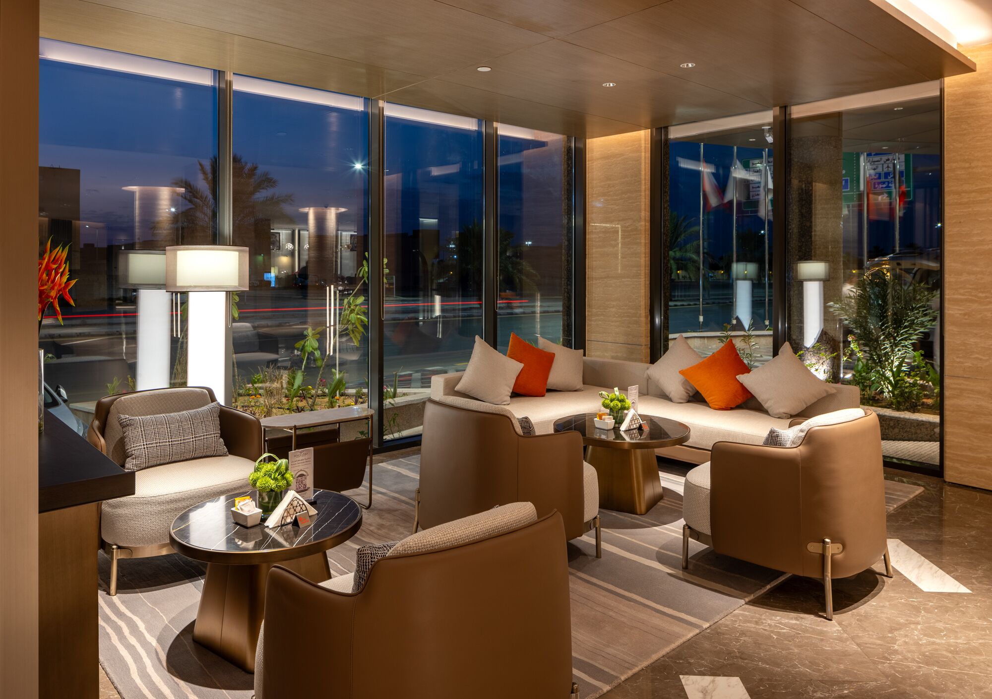 A modern hotel lobby with a sofa set, armchairs, a coffee table, and a lamp. Large windows offer a view of the cityscape at night.