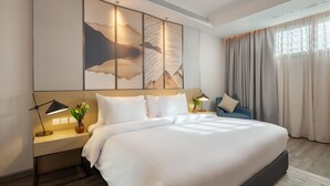 Una habitación de hotel moderna con una cama grande, una mesita de noche con una lámpara, un florero con flores y vistas al exterior a través de cortinas translúcidas.