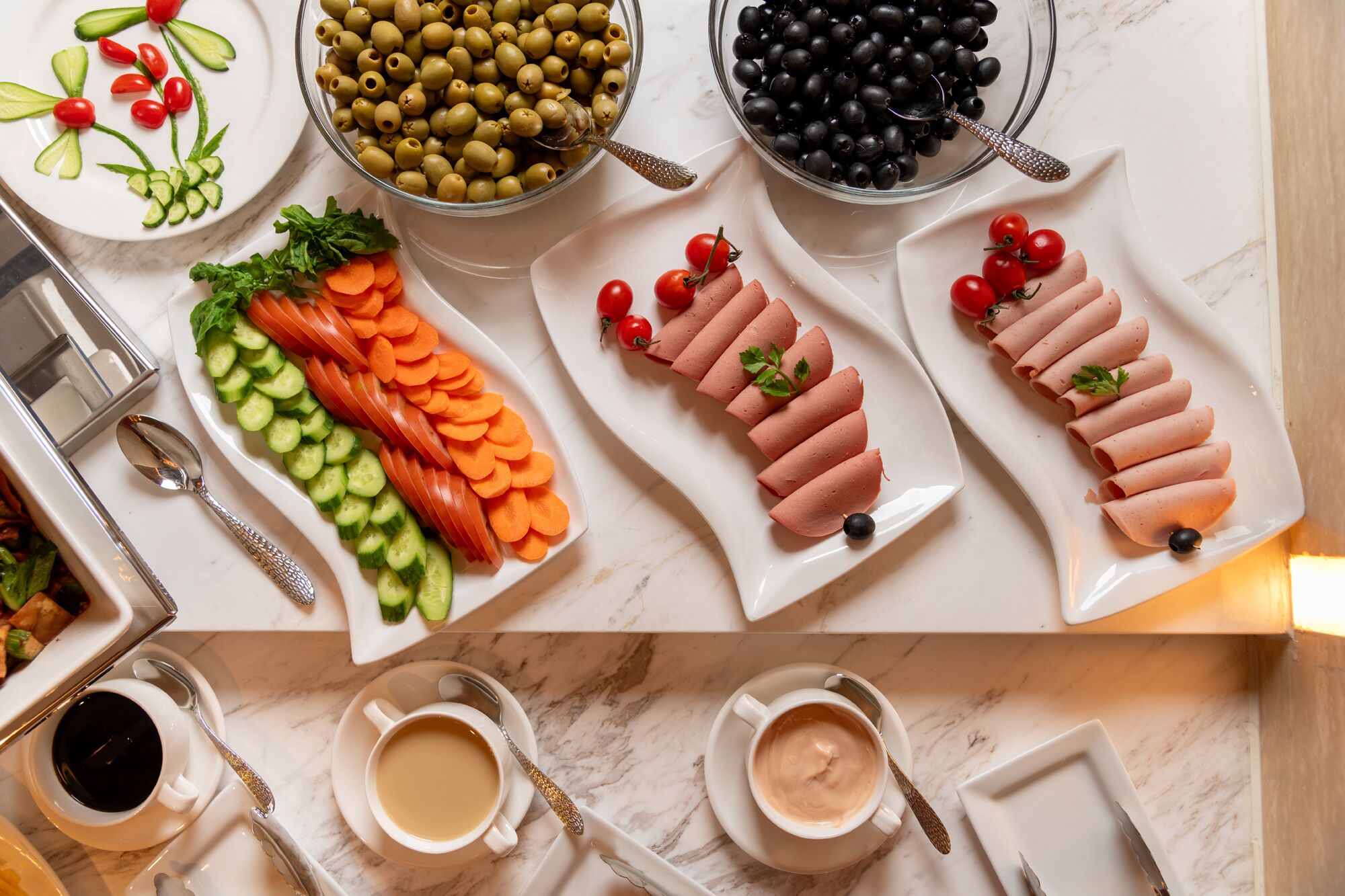 A table with various food items and drinks, including sliced cucumbers, carrots, cherry tomatoes, olives, and blackberries, as well as two cups of coffee and a plate of sliced meat.