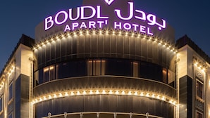 El hotel-apartamento Boudl Jagh con un llamativo letrero iluminado, varias banderas y una entrada bien iluminada.