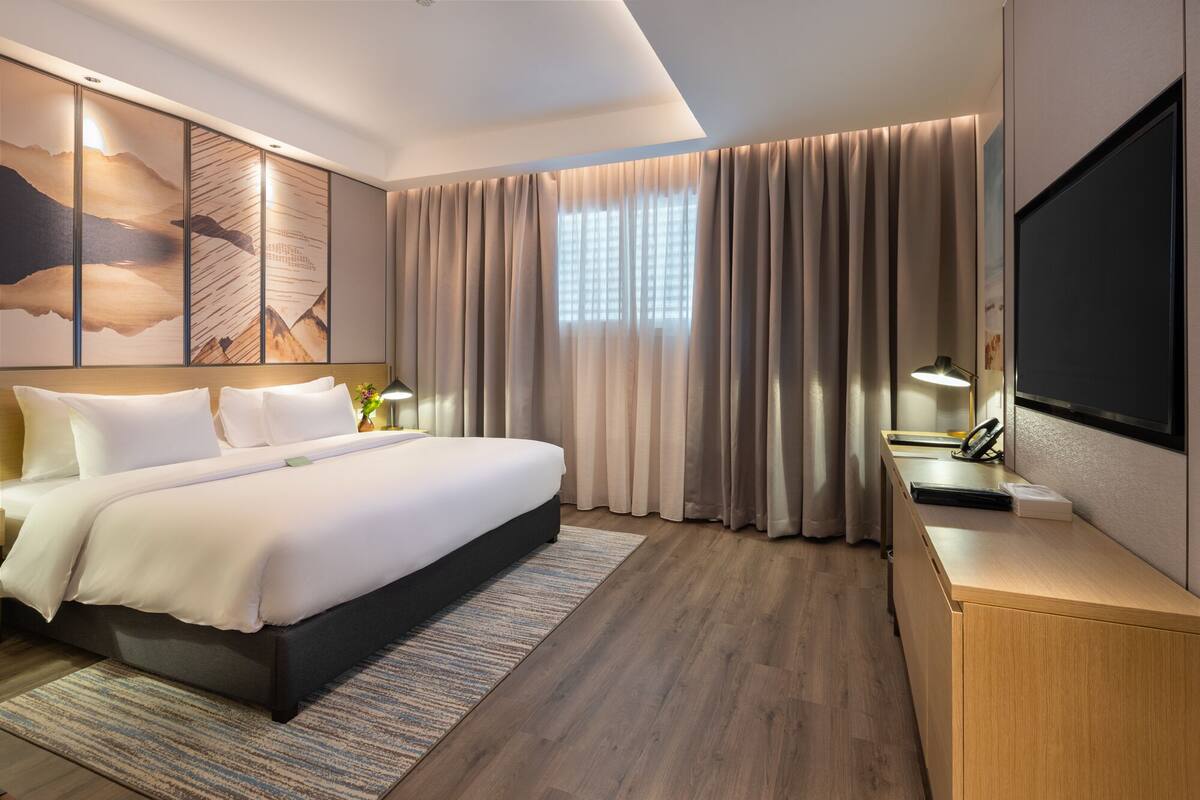 Una habitación de hotel moderna con una cama grande, un escritorio con televisor de pantalla plana y un ventanal con cortinas.